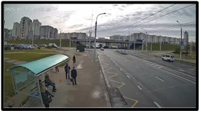 Overview (OV) camera wide shot of a bus stop and pedestrians beside a multi-lane road with apartment buildings in the background