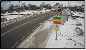 TL camera view of a snowy road approaching a railway crossing with a warning sign showing a national flag and a barrier arm visible ahead
