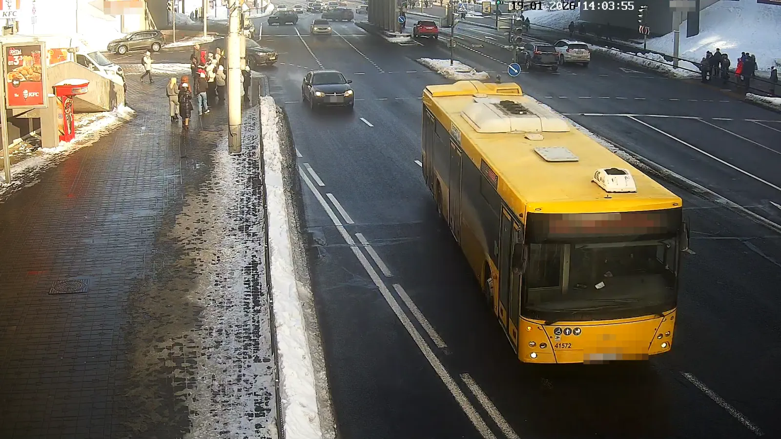 OV camera wide shot of a city street with a yellow bus, pedestrians waiting at a stop, and traffic in a snowy setting