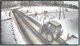 OV camera wide shot of a tractor and workers maintaining a snowy railway crossing with barrier arms and warning lights