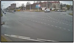 OV camera wide shot of a wide multi-lane intersection with sparse traffic and roadside buildings in view