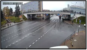 OV camera wide shot of an intersection beneath an overpass with a vehicle and pedestrians visible on a wet road