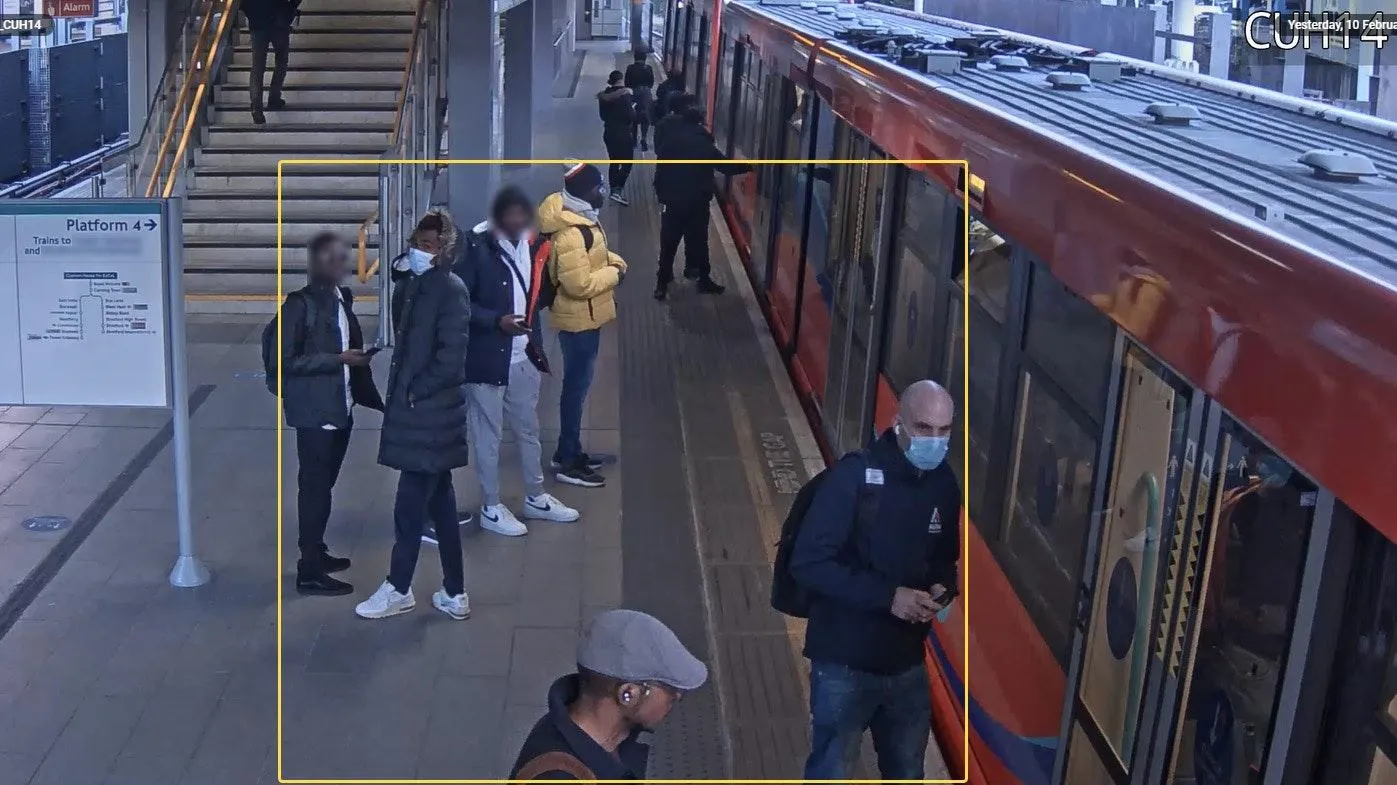 Railway platform camera view with a yellow bounding box drawn around a group of waiting passengers
